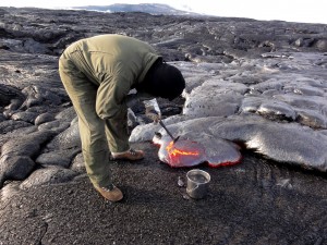 An HVO geologist collects a molten lava sample for chemical analysis, scooping up a bit with the rock hammer to then drop in the water bucket to quench it. Puʻu ʻŌʻō is visible in the distance. Photo taken Wednesday, December 30, 2015 courtesy of USGS/HVO