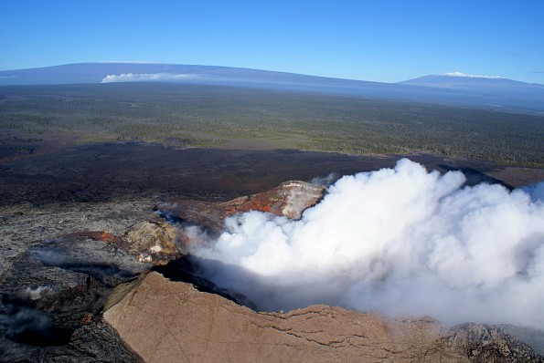 During a kona wind, fume from Pu‘u ‘Ō‘ō (foreground) and Halema‘uma‘u Crater (background), both on Kīlauea, blows northward, with towering Mauna Loa and Mauna Kea on the horizon. USGS photo.
