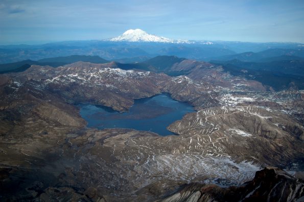 Aerial view of Spirit Lake (center) looking north from above the crater of Mount St. Helens. The lake’s outlet to the west (left) was blocked by the 1980 landslide, which required a new engineered outlet to maintain the lake at a safe level. A tunnel was drilled through a bedrock ridge on west side of lake in 1984-1985. Glacier covered Mount Rainier in distance. USGS photo by M. Logan, 2005.