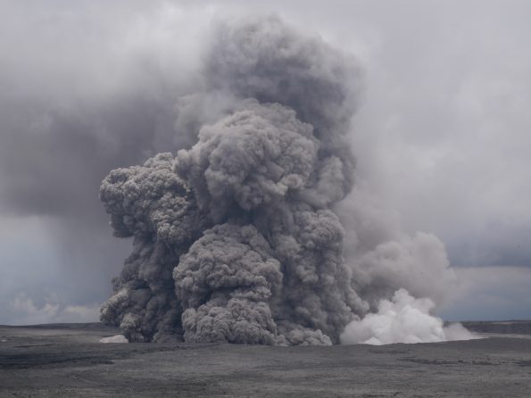 Ash rises above Halemaʻumaʻu within Kīlauea’s summit caldera in this May 27, 2018, telephoto image from near Volcano House Hotel in Hawaiʻi Volcanoes National Park. By the time Kīlauea’s summit collapse events ended on August 2, Halema‘uma‘u was 2.5 km (1.5 mi) wide and 500 m (1600 ft) deep; prior to the 2018 collapses, it was about 1 km (0.5 mi) wide and 85 m (about 280 ft) deep. A segment of a long-closed Park trail is visible winding across the caldera floor (lower left). USGS photo by K. Anderson.